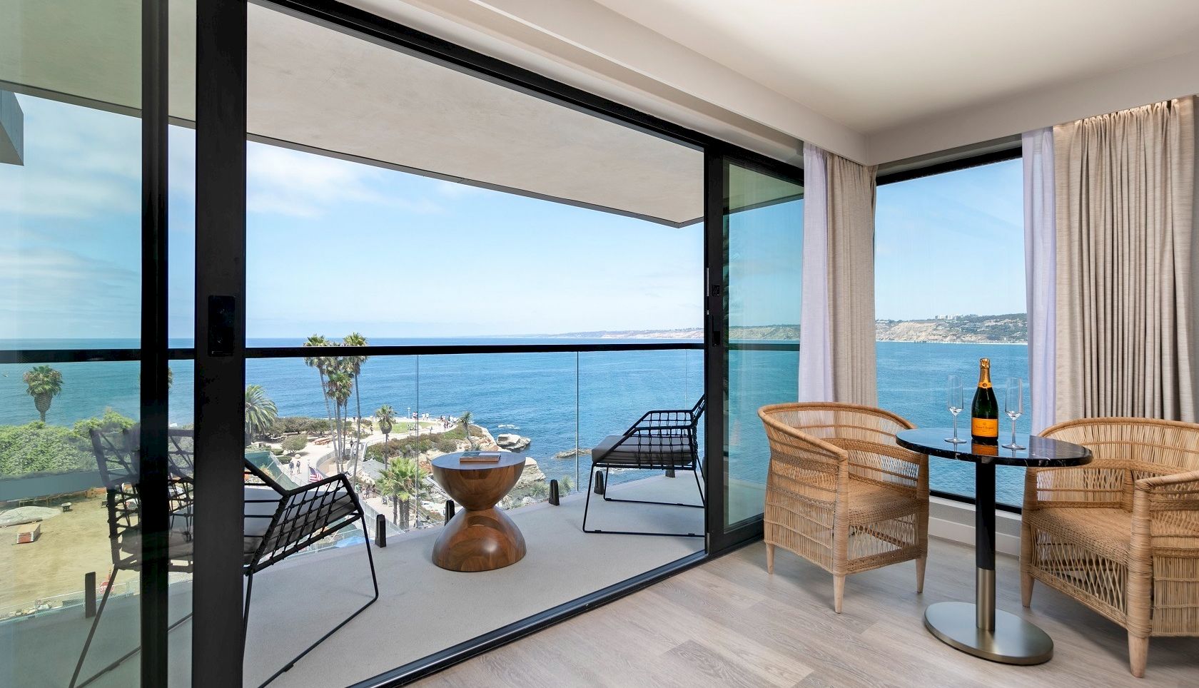 Modern balcony with wicker chairs and a table, overlooking the ocean under a bright sky.