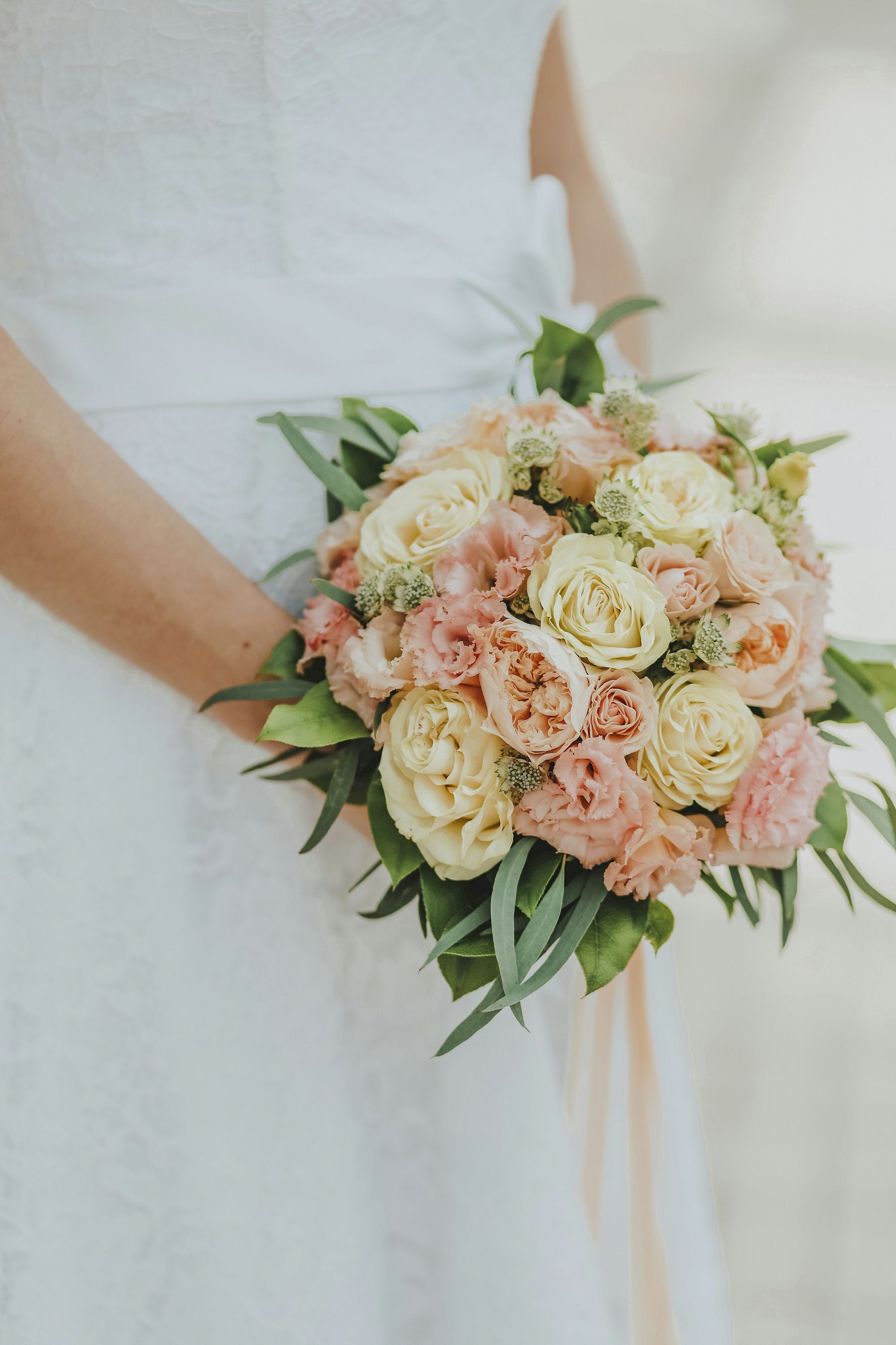 A pair of hands holds a soft pastel bridal bouquet of cream and blush roses, baby&rsquo;s breath, and lush green leaves.