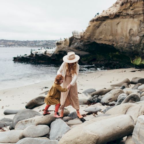 A woman in a hat holds hands with a child on a rocky beach by the ocean and cliffs.