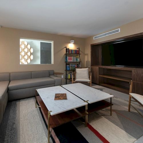 Modern living room with a gray sectional, marble coffee table, wall art, bookshelves, and a large flat-screen TV.