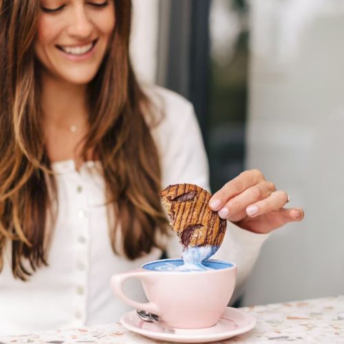 Smiling woman holds a grilled pastry over a cup, pouring filling into tea/coffee during breakfast.