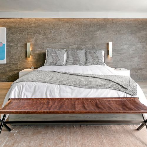 A modern bedroom with a white bed, patterned pillows, warm wall lights, and a textured stone wall background.