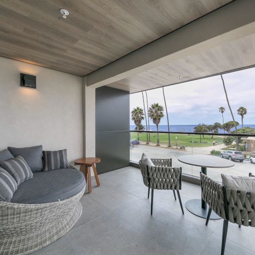A modern patio living area with sofas and dining chairs, glass railing, and a view of palm trees and the ocean.