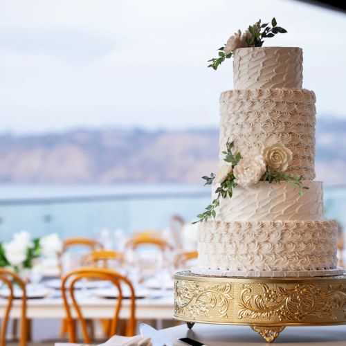 A tiered white wedding cake with floral decorations sits on a gold stand beside a seaside table setting.