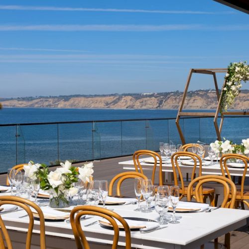Sunlit seaside terrace dining with white table settings, golden chairs, and a floral wedding arch overlooking the ocean.