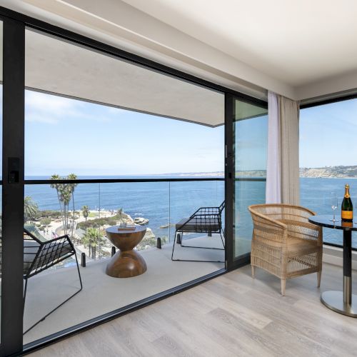 A seaside balcony with rattan chairs, a small table with champagne, and big sliding doors opening to the blue ocean.