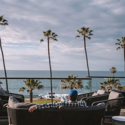 A man relaxes on a seaside patio with palm trees, lounge chairs, and a cloudy ocean horizon behind him.
