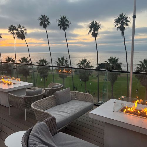 Oceanfront patio at sunset with palm trees and glass railings, plus glowing fire pits on tables.