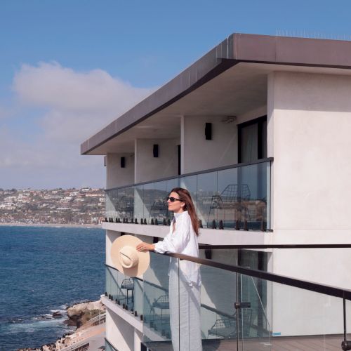 A woman in a white dress stands on a cruise ship balcony, holding a hat, overlooking the ocean and a coastal town.