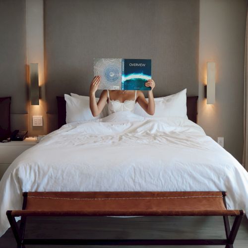 In a hotel room, a woman lies on a bed holding up a magazine to cover her face against a gray wall.