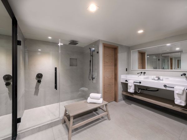 A modern, spacious bathroom with a glass-enclosed shower, a long double vanity, large mirrors, bright lighting, and a small wooden bench with towels.