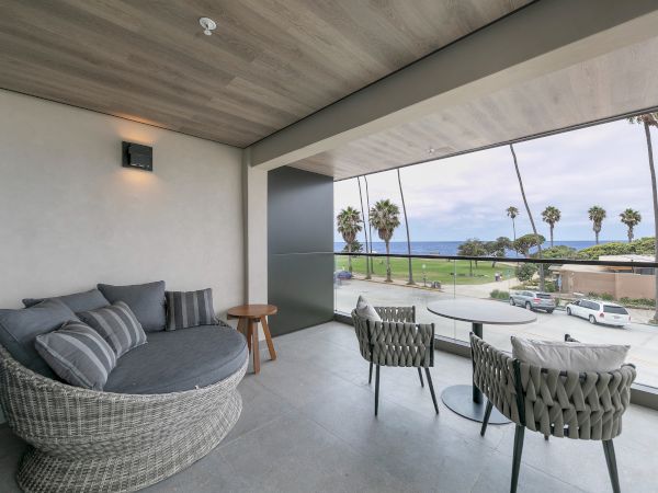 Cozy covered balcony with woven sofa, striped cushions, round table, and wicker chairs overlooking a sunny street and palm trees by the beach.