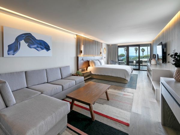 A modern hotel suite with a gray sectional, wooden coffee table, and king bed; balcony view, contemporary art, and clean minimal decor.
