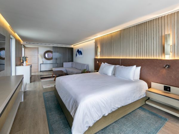 A modern hotel room with a large bed, soft lighting, a seating area, and a slim desk; airy decor in neutral tones with a blue rug.