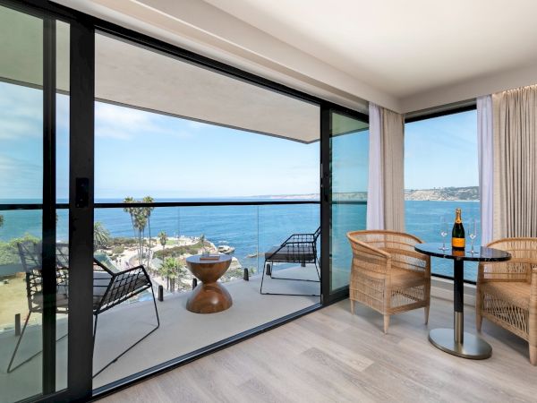 A modern hotel room with a balcony overlooking the ocean; sliding glass doors, wicker seating, and a small table with chairs.