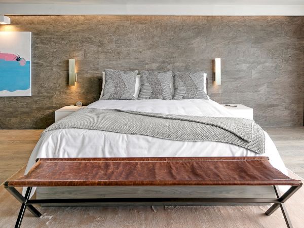 A modern minimalist bedroom with a large bed, gray bedding, wooden bench at the foot, and textured concrete wall lighting&mdash;tiled, sleek, serene.