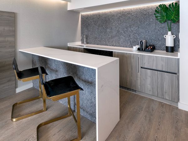 Modern kitchenette with a gray-blue backsplash, floating cabinets, a white island, two slim chairs, and warm wood tones. End sentence.