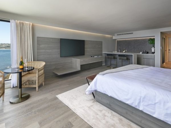 A modern hotel room with a wall-mounted TV, a floating dresser, a bed, a kitchenette, a small dining table by the window, and a sea view.