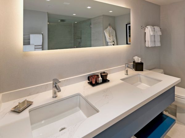 A modern bathroom with a long white double sink, a large backlit mirror, light-gray walls, and open shelf storage beneath the countertop.