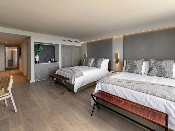 A modern hotel room featuring two large beds, a desk, seating area, ambient lighting, and a hallway leading to other spaces.