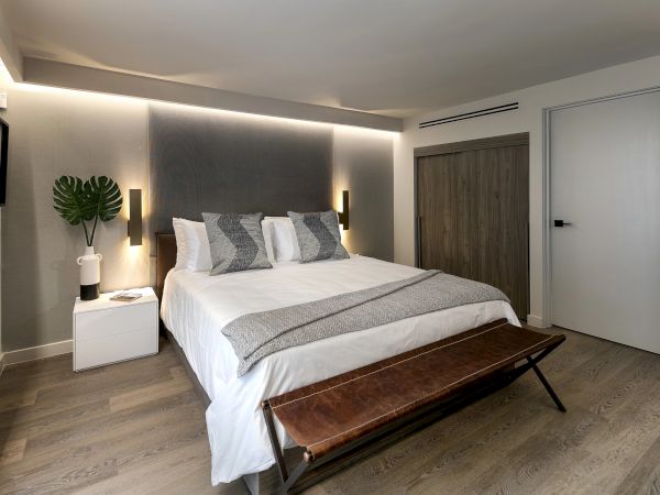 A modern hotel bedroom with a comfy queen bed, gray and white decor, wall sconces, a bench at the foot, a white nightstand, and a wooden door in a bright, cozy space.
