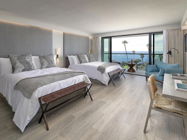 A bright hotel room with two queen beds, soft gray headboards, modern decor, a seating area, and a balcony view of the ocean.