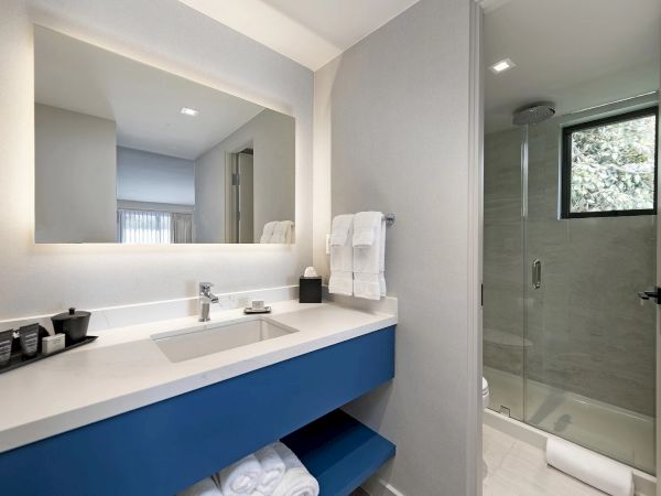 A modern bathroom with a double sink vanity, blue open shelf, large backlit mirror, and a glass-walled shower.