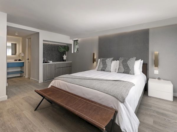 A modern hotel-style bedroom with a large bed, gray headboard, two nightstands, bench at foot, airy light wood floors, and a small open kitchenette area in the background.