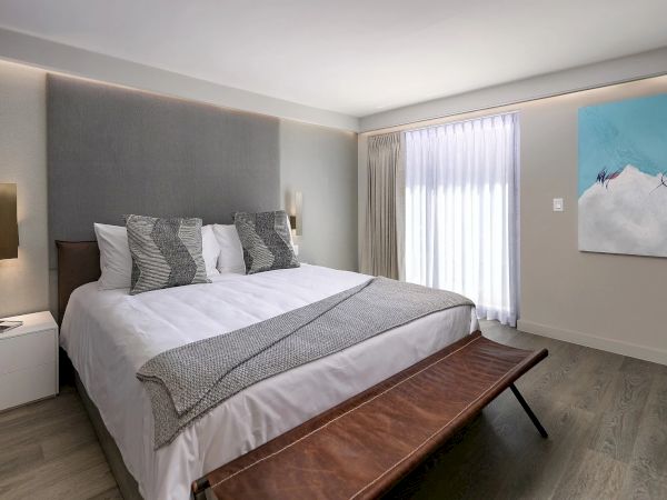 A modern hotel room with a large bed, gray upholstered headboard, white bedding with gray accents, two bedside lamps, a wooden bench at the foot, light wood floors, and a blue abstract wall art piece by the window.