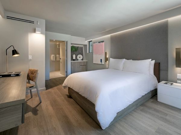 A modern hotel room with a large bed, work desk, flat-screen, and soft grey/white decor; en suite bathroom visible through open door.