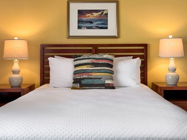 A cozy hotel-style bed with white bedding, a patterned lumbar pillow, two matching nightstands, and warm bedside lamps framing a framed sunset picture.
