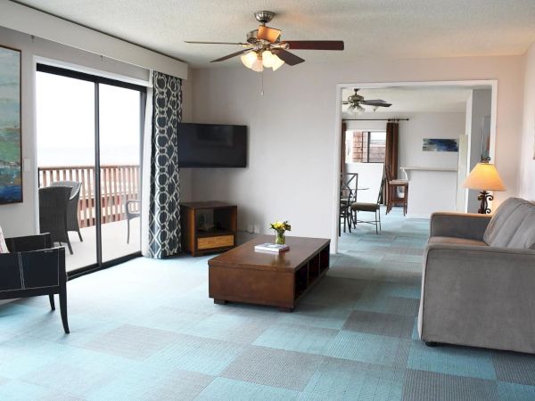 A bright hotel suite living room with a sofa, coffee table, TV, sliding glass door to a balcony, and a doorway to another seating area.