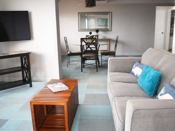 A modern living room with a gray sofa, blue throw pillows, a wooden side table, and a dining area in the back featuring a table and chairs.