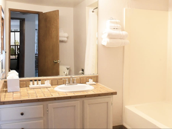 A bathroom with a single sink vanity, beige tiled countertop, mirror, towel rack with white towels, and a bathtub/shower combo.
