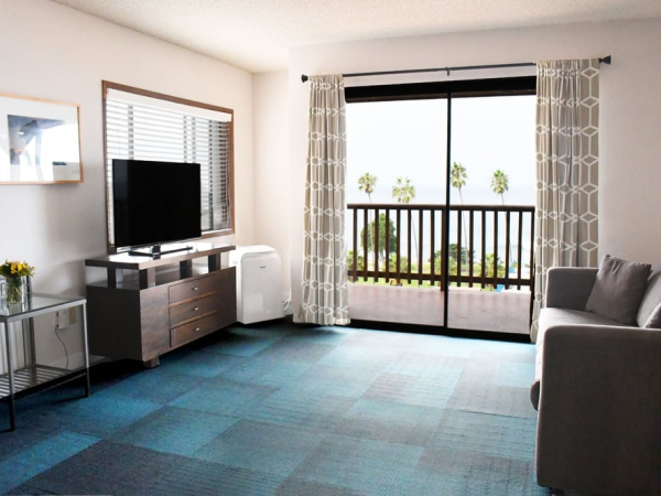 A bright hotel living area with a TV on a console, a sofa, sliding glass door to a balcony, and blue patterned carpet. End.