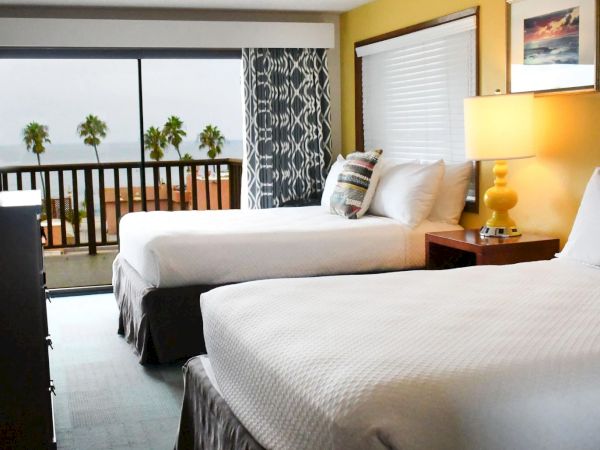 A hotel room with two white beds, a balcony view of palm trees, yellow accent wall, and a nightstand lamp&mdash;bright, airy, and inviting.