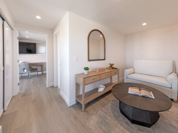 A bright, modern living area with a sofa, oval coffee table, rug, and a wooden console with a mirror; hallway leads to a kitchen.