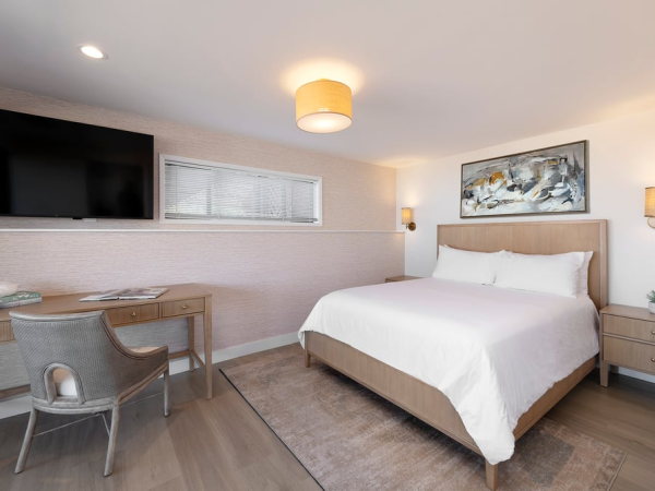 A modern hotel room with a large bed, nightstands, a wall-mounted TV, a desk and chair, a rug, soft lighting, and light wood tones, all tidy and inviting.