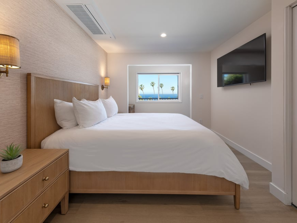 A bright hotel room with a king bed, nightstands, wall sconces, a wall-mounted TV, and a window view of palm trees and ocean, modern and tidy.