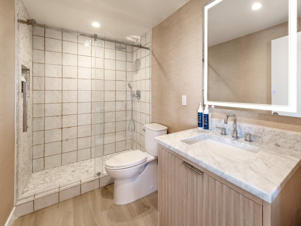 Modern bathroom with a tiled walk-in shower, toilet, and marble countertop vanity with a backlit mirror, neutral tones throughout.