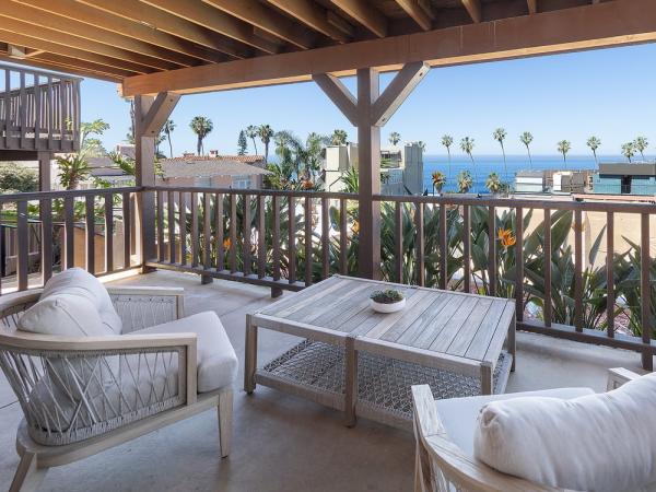 A sunny balcony with light wicker furniture, a wooden table, and white cushions, overlooking palm trees and the ocean beyond.