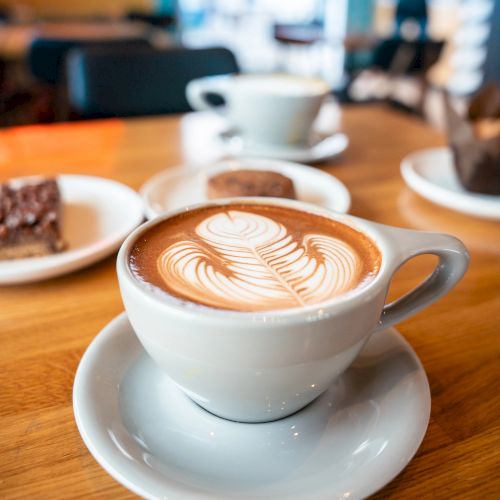 A latte with latte art in a white cup on a wooden table, coffee shop scene, with a plate of dessert and another cup in the background.