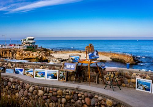 An outdoor seafront gallery with framed photos displayed along a stone wall, a small viewing deck by the sea, and a clear blue sky over calm water.