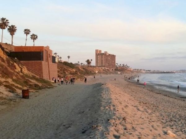 A sunny beachfront with sandy shore, palm trees, a few people walking, and buildings along the coast fading into the distance.