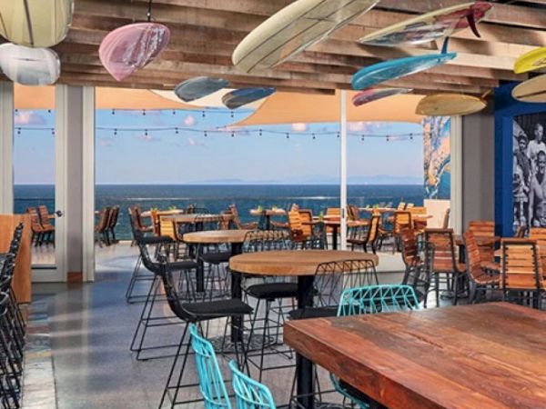 A seaside patio restaurant with wooden tables and chairs, overlooking the ocean, featuring colorful hanging ornaments above it.