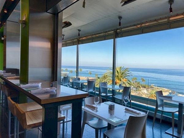 A seaside restaurant terrace with neatly set tables and chairs, ocean view through large glass windows.