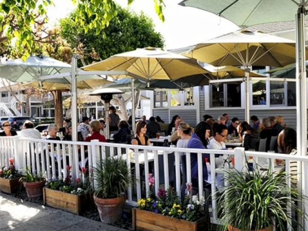 A bustling outdoor restaurant patio with diners seated under yellow and green umbrellas, surrounded by potted plants and trees.