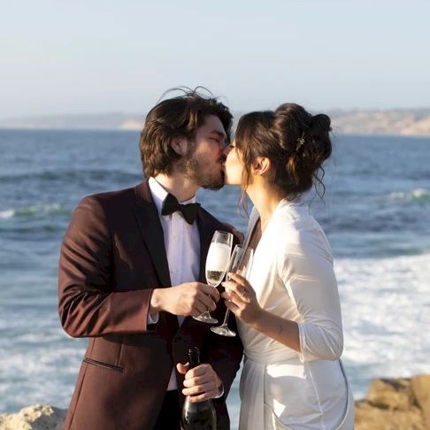 A couple on a seaside beach kisses while toasting with champagne glasses in formal attire.