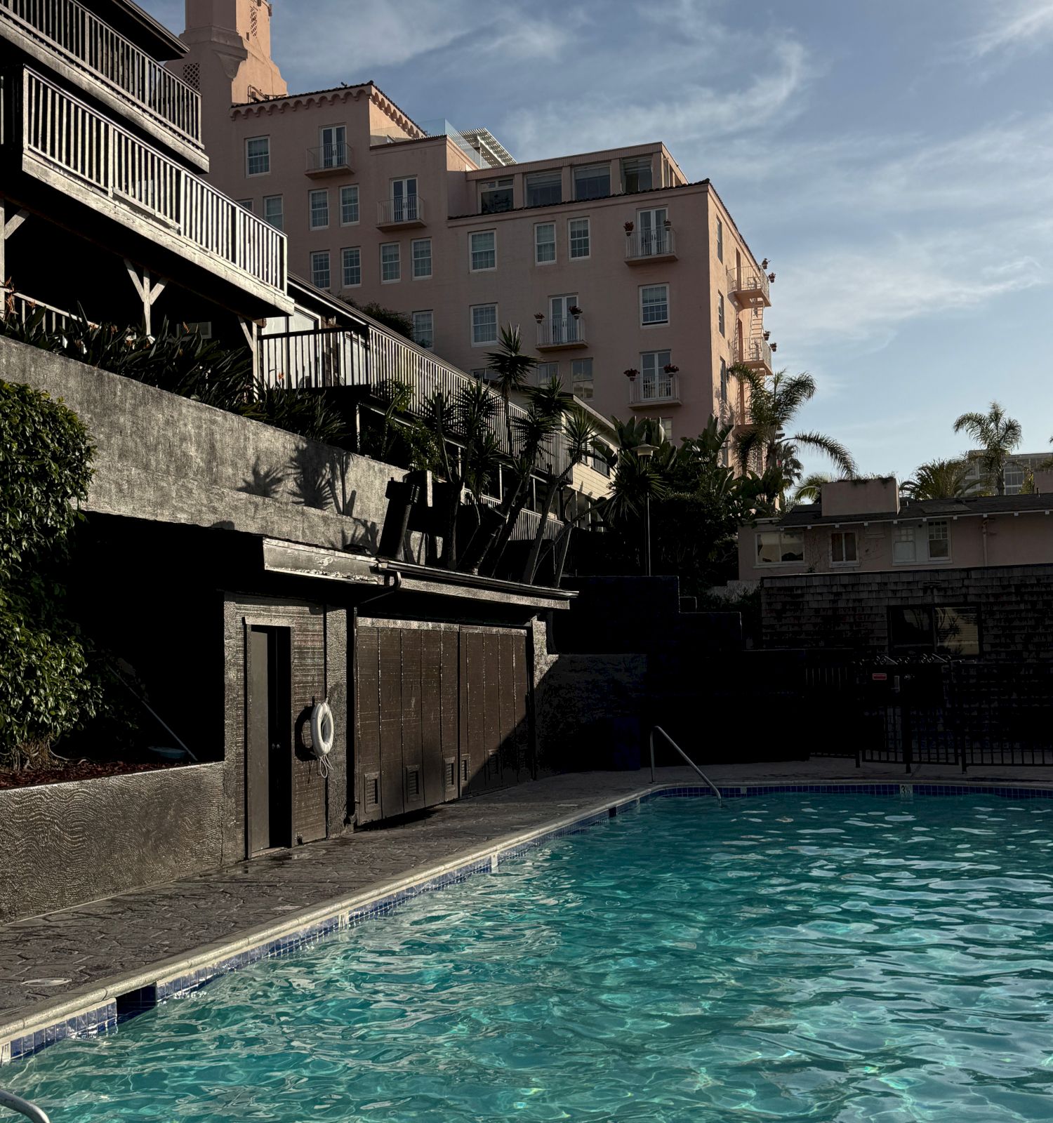 Outdoor hotel courtyard pool with palm trees and a beige building rising to a tall tower under cloudy sky.