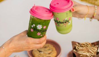 Two people hold up matching green drinks with pink lids over a table of pastries and cookies, in warm daylight.